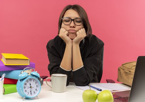 手疲惫的年轻女生戴着眼镜坐在办公桌上用大学工具把双手放在下巴下闭着眼睛做作业 粉红的背景隔开关工具大学