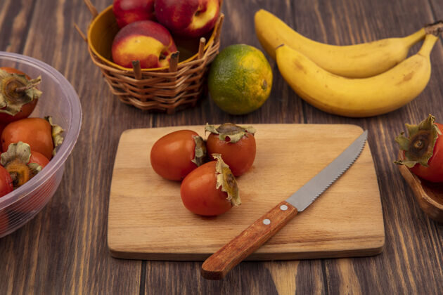 视野木制菜板上甜柿的俯视图 木桶上放着桃子和小刀 木制背景上隔离着橘子和香蕉食物顶部柿子