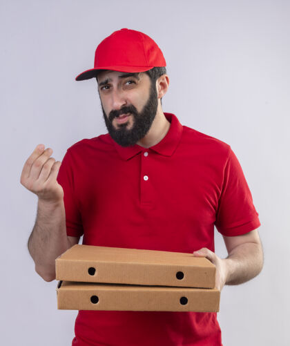 拿着年轻英俊的白人送货员身穿红色制服 头戴鸭舌帽 手里拿着比萨饼盒 指着钞票 看着隔离在白色背景上的摄像机手势盒子披萨