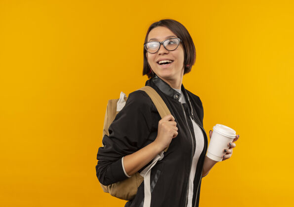穿着快乐的年轻学生女孩戴着眼镜 背着书包站在侧视图后面看 手里拿着咖啡杯孤立在橙色喜悦视图眼镜