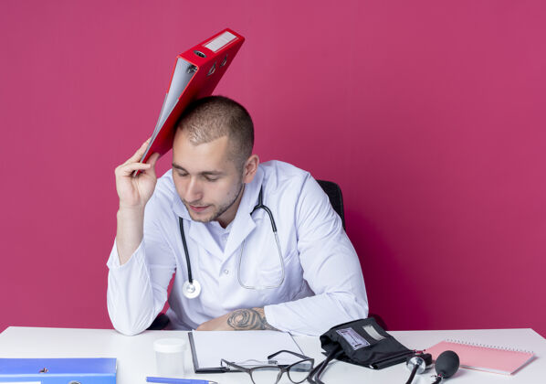 头疲惫的年轻男性医生穿着医用长袍和听诊器坐在办公桌旁 手里拿着工作工具 拿着文件夹 闭着眼睛 用它摸着头 隔离在粉色背景上男性持有封闭