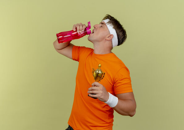 优胜者年轻帅气的运动型男人戴着头带和腕带 手里拿着优胜者杯 喝着橄榄绿背景上孤立的水瓶里的水 还有复制空间年轻头带运动