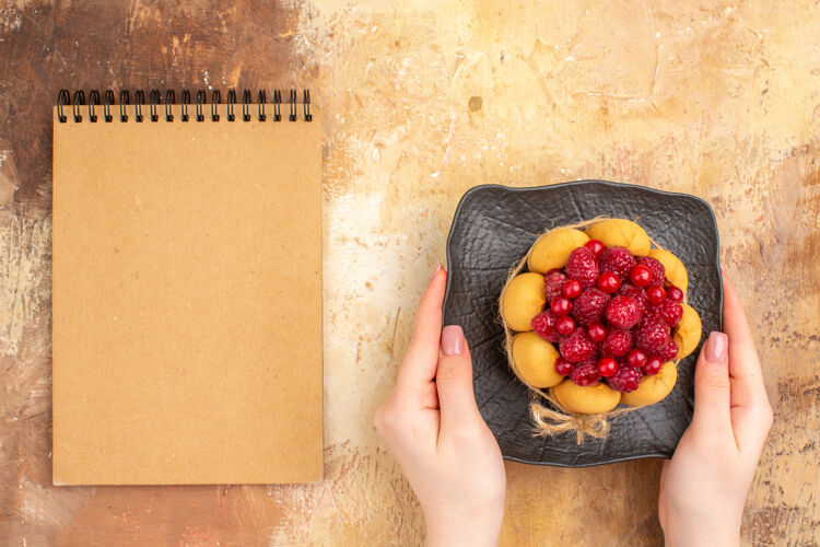 浆果手拿着刚烤好的礼品蛋糕和笔记本放在一个棕色的盘子上 放在五颜六色的桌子上保持混合食物
