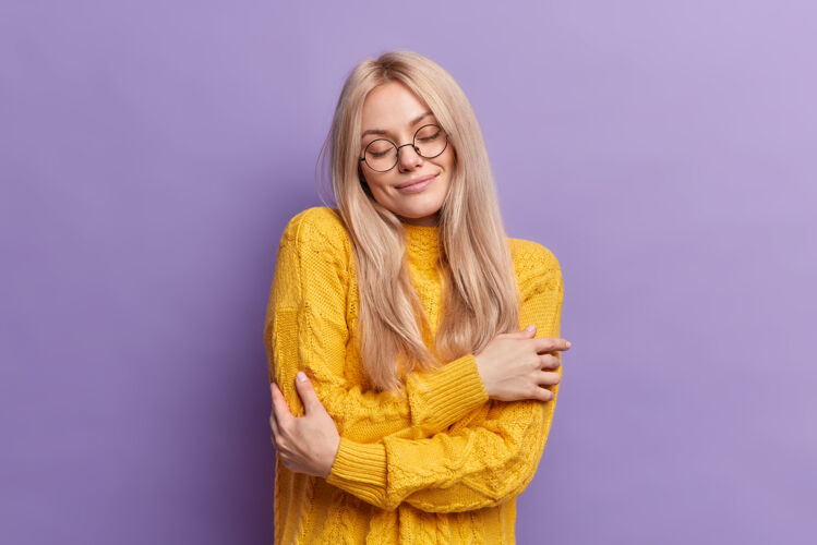 风格可爱满意的金发女人拥抱自己感觉舒服穿着柔软的黄色毛衣闭上眼睛对着鲜艳的紫色墙壁摆姿势眼镜眼镜乐趣