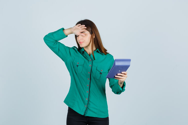 成人年轻女士拿着计算器 手放在额头上 穿着绿色衬衫 看上去很沮丧前视图女尖叫保持