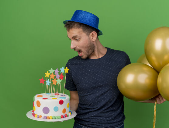 生日戴着蓝色派对帽的高加索帅哥兴奋地拿着氦气球 看着隔离在绿色背景上的生日蛋糕绿色帽子蓝色