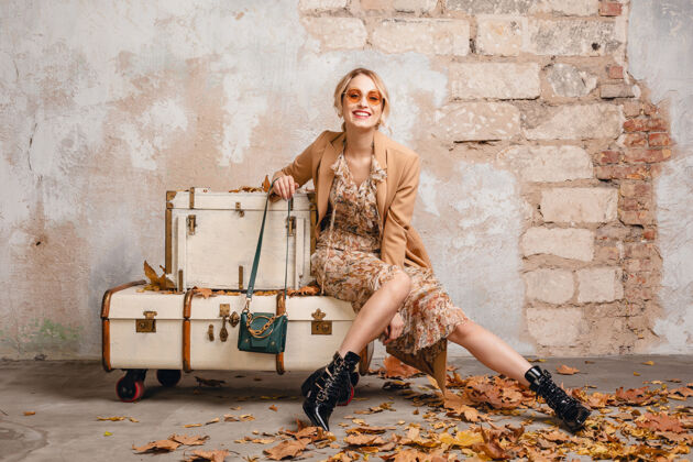 时尚在街上 穿着米色外套的金发美女坐在手提箱上对着墙女士时尚城市