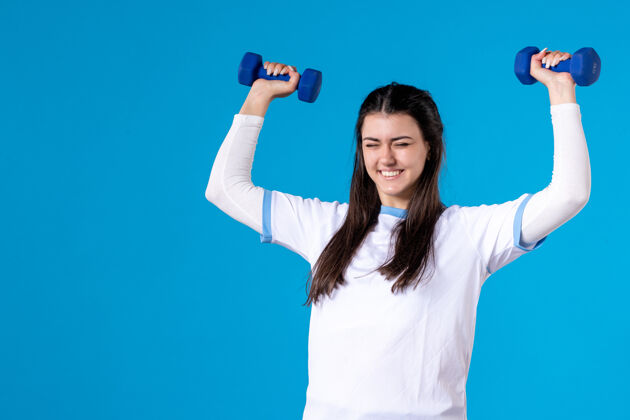 蓝色正面图：年轻女子手持蓝色哑铃站在蓝色墙上身体成人健身