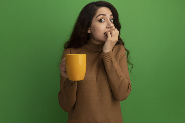 杯子关心的年轻漂亮的女孩拿着一杯茶咬着被隔离在绿墙上的钉子女孩关心茶