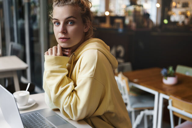 金发年轻漂亮的女孩坐在咖啡馆里拿着笔记本电脑女学生在咖啡馆学习聊天笔记本电脑电脑