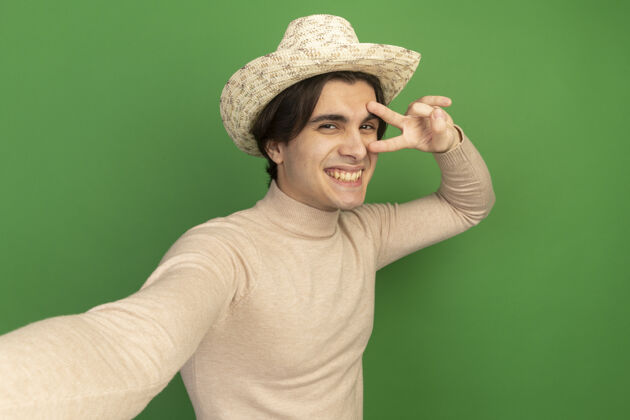 表演带着微笑的年轻帅哥戴着帽子在绿色的墙上显示出和平的姿态和平帽子年轻
