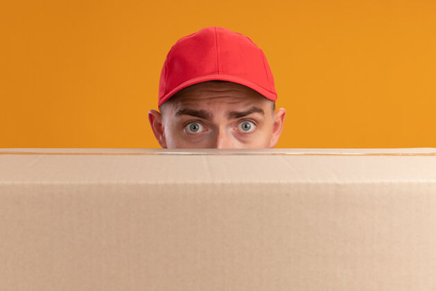 封面惊讶的年轻送货员穿着制服 戴着帽子 脸上有一个大盒子 隔离在橙色的墙上制服帽子男人