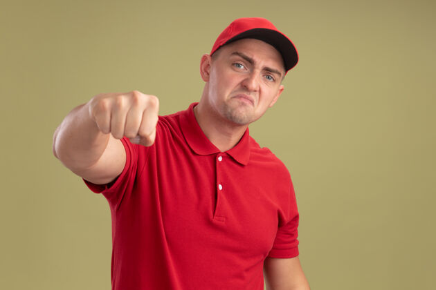 年轻愤怒的年轻送货员穿着制服 戴着帽子 在橄榄绿的墙上对着镜头伸出拳头穿举行男人