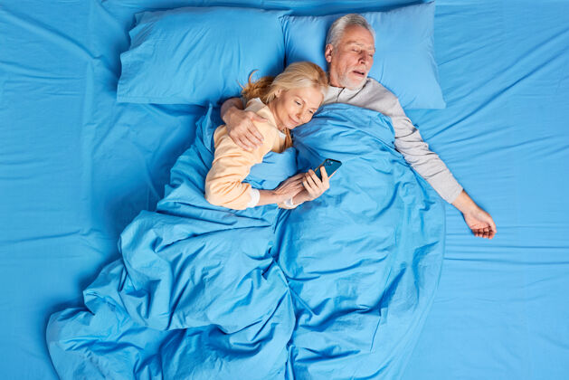 在一起沉迷于现代科技的女人在床上用手机在睡前接受熟睡丈夫的拥抱上网中年夫妇在舒适的卧室里休息男人睡觉拥抱