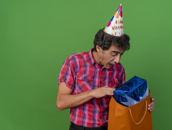 副本令人印象深刻的中年白种人聚会男子戴着生日帽看里面的纸袋与礼品包隔离在绿色背景与复制空间印象绿色聚会