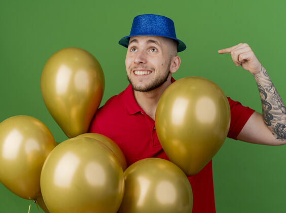 指着面带微笑的年轻英俊的斯拉夫党人戴着党的帽子站在气球后面看着一边指着孤立在绿色背景上的气球气球斯拉夫人站着
