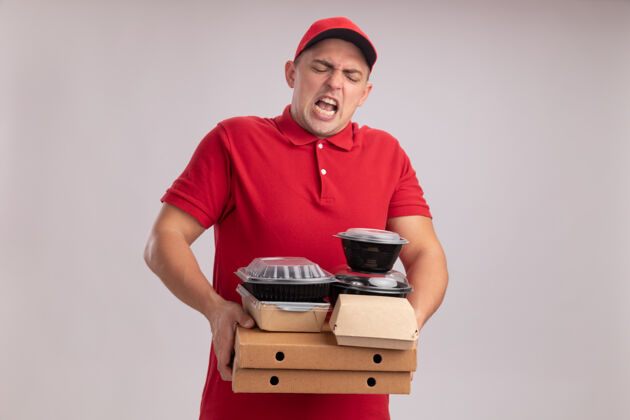 穿紧张的年轻送货员穿着制服 戴着帽子 把食物容器放在隔离在白墙上的比萨饼盒上帽子拿着盒子