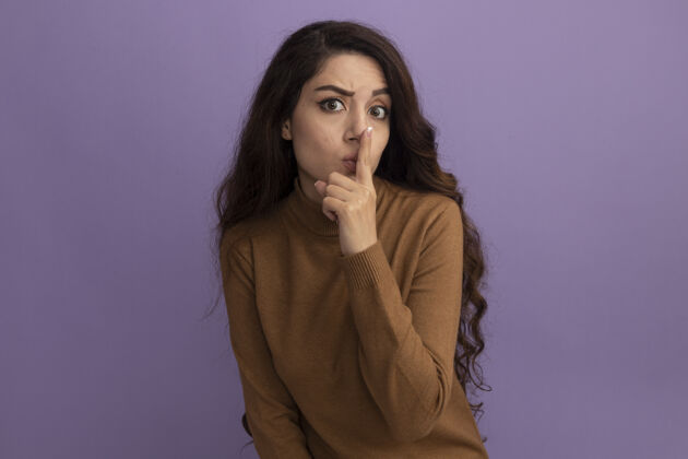 手势穿着棕色高领毛衣的年轻漂亮女孩在紫色的墙上表现出沉默的姿态沉默严格高领毛衣