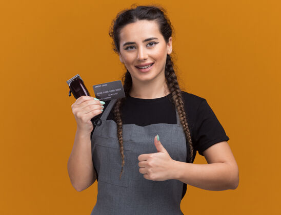 年轻微笑着的年轻女理发师穿着制服拿着信用卡和理发器在橙色的墙上孤立地竖起大拇指持有女信用卡