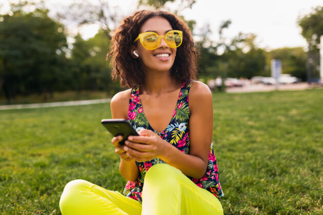 人年轻时尚微笑的黑人女子使用智能手机 通过无线耳机听音乐 在公园里玩得很开心混血儿年轻黄色