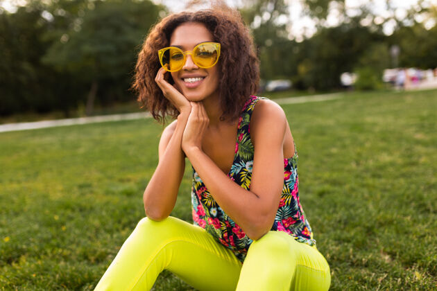 女孩年轻时尚的黑人女子在公园里尽情享受夏日的时尚风格 五颜六色的时髦装扮 戴着黄色太阳镜坐在草地上卷曲五颜六色肖像