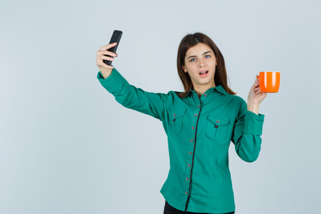 采取一位年轻的女士端着一杯橙黄色的茶 穿着衬衫拿着手机自拍 看上去很自信正面图衬衫焦点手机