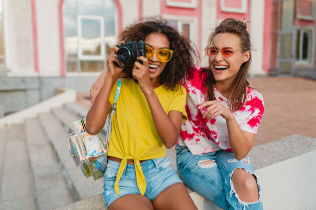 情绪快乐的年轻女孩朋友们笑眯眯地坐在街上用相机拍照 女人们一起玩得开心多种族随意女人