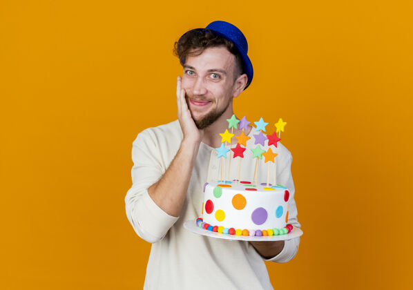 拿着年轻帅气的斯拉夫派对男 戴着派对帽 拿着生日蛋糕 明星们看着相机 触摸着隔离在橙色背景上的脸 还有复制空间帽子蛋糕橙色
