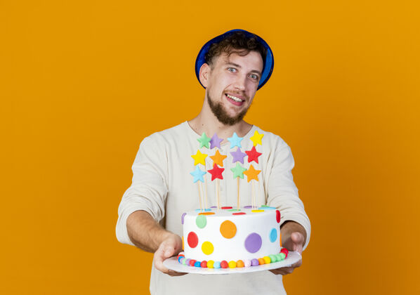 生日面带微笑的年轻帅哥斯拉夫党家伙戴着党帽子伸展生日蛋糕与星星和看相机孤立在橙色背景与复制空间伸展太空斯拉夫