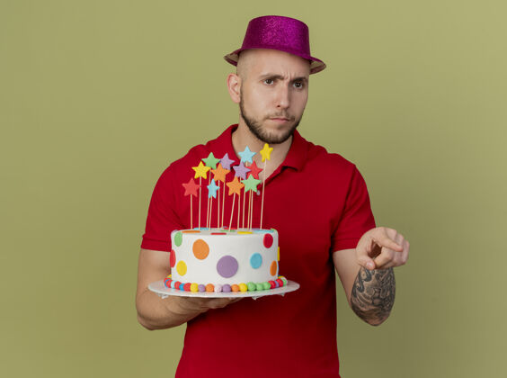 年轻皱眉年轻英俊的斯拉夫党人戴着党的帽子拿着生日蛋糕看着并指着一边孤立的橄榄绿背景与复制空间复制皱眉生日