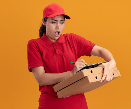 什么的一个穿着制服 戴着帽子的年轻送货女孩被吓坏了 她在剪贴板上写着什么 就在隔离在橙色墙上的比萨饼盒上害怕剪贴板帽子