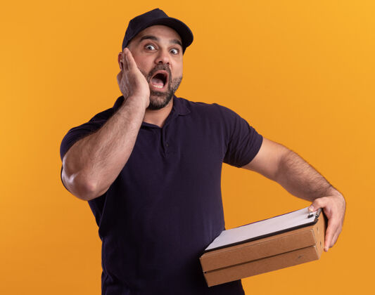 男人吓坏了的中年送货员 穿着制服 戴着帽子 拿着披萨盒的剪贴板 手放在隔离在黄色墙上的脸颊上拿着披萨害怕