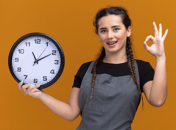 钟微笑着的年轻女理发师穿着制服 拿着挂在橙色墙上的挂钟 显示出良好的姿态好的年轻的女的