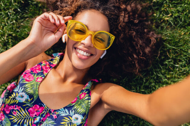 黄色年轻时尚的黑人女子 戴着无线耳机听音乐 躺在公园的草地上玩得很开心 用手机相机拍自拍 夏日时尚风格多彩的时髦装扮 俯瞰风景眼镜欢呼太阳镜