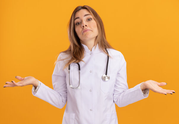 困惑困惑的年轻女医生穿着医用长袍 听诊器摊开双手隔离在黄色的墙上传播年轻穿