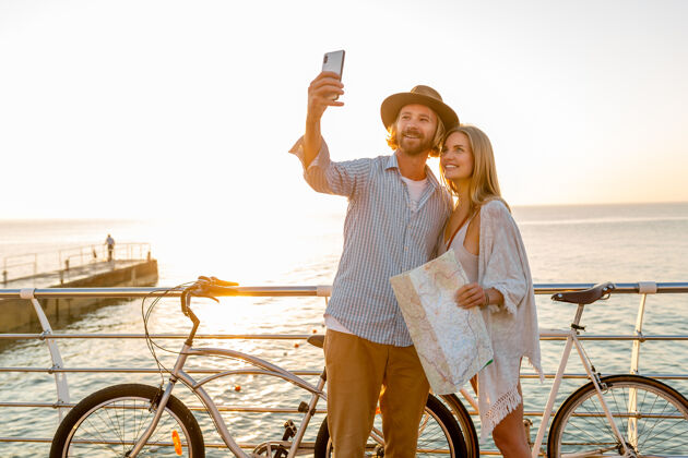 微笑年轻迷人的笑容可掬的快乐男女骑着自行车在手机摄像头上自拍 夕阳下海边的浪漫情侣 波西米亚时髦的装扮 朋友们在一起玩得开心朋友阳光日落
