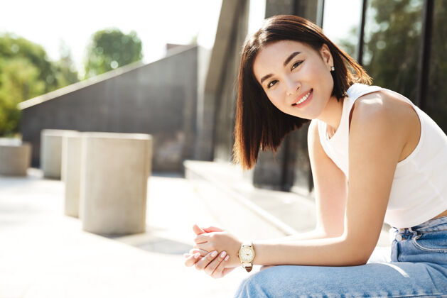 花园年轻的亚洲女人坐在大楼附近的长凳上 开心地对着镜头微笑无忧无虑乐趣城市