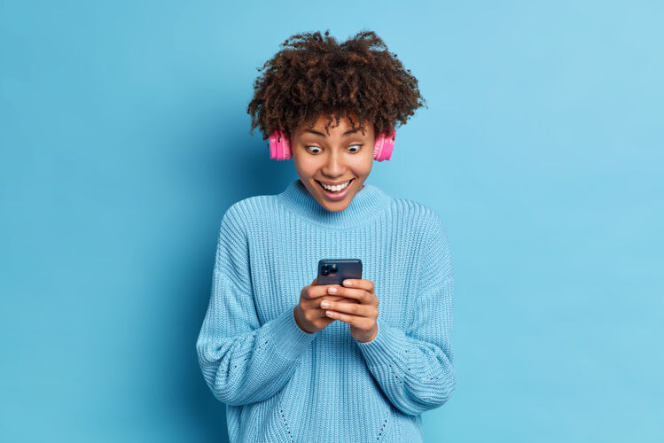 套头衫一张非洲裔美国少女凝视着智能手机屏幕的照片读着惊人的新闻戴着粉色立体声耳机穿着针织毛衣听音乐高兴卷发正面