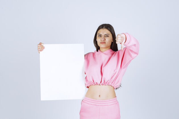 休闲穿着粉色睡衣的女孩拿着一块空白的正方形展示板 拇指朝下人类人体模特工人