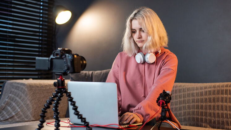 女孩年轻的内容创作者金发女孩戴着耳机在桌上的笔记本电脑上工作 带着相机女人科技女性