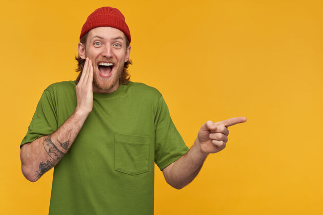 胡须笑容可掬的金发胡子 穿着绿色t恤和红色小珠子 身上有纹身手指指向右边的复制空间 隔离在黄色的墙上年轻触觉情绪