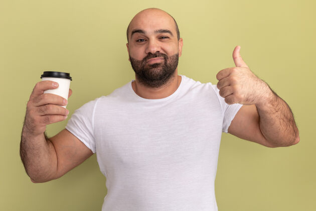 正面胡子男人穿着白色t恤 拿着咖啡杯快乐而积极地站在绿色的墙上伙计杯子咖啡