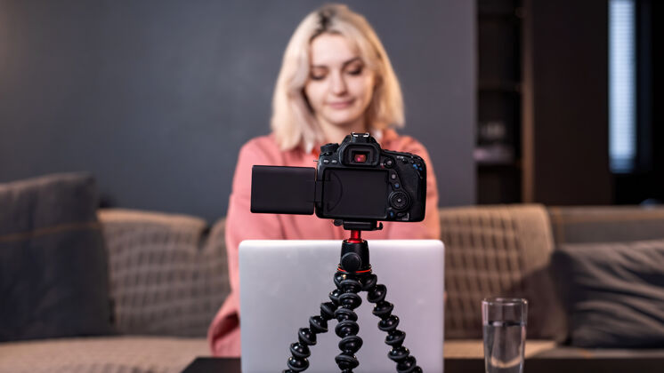屏幕年轻的内容创作者金发女孩在桌上的笔记本电脑上用三脚架上的相机拍摄自己在家工作录制视频日志鼠标工作女性