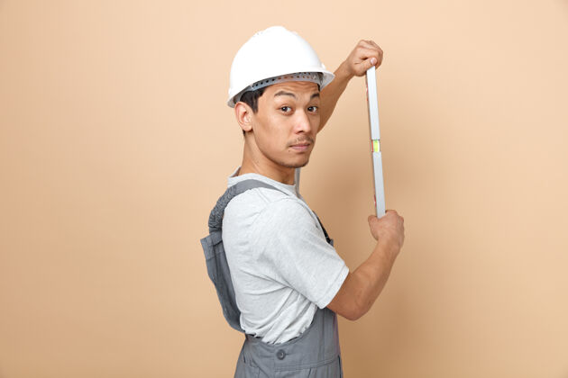 安全年轻的建筑工人戴着安全帽 穿着制服 手持水平尺站在纵断面图上复制建筑制服