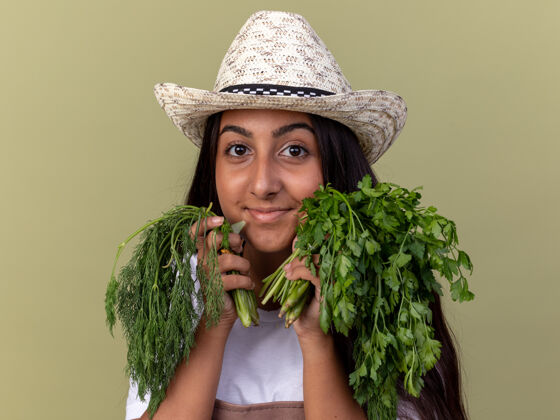站戴着围裙和夏帽的快乐的年轻园丁女孩站在绿色的墙上 手里拿着新鲜的草药 脸上带着微笑围裙帽子花园