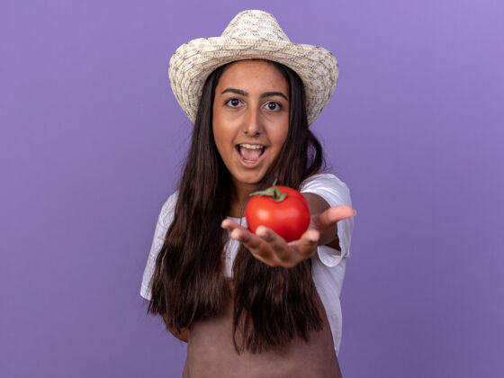 微笑戴着围裙和夏帽的快乐的年轻园丁女孩站在紫色的墙上 脸上带着微笑 展示着新鲜的西红柿帽子站着围裙