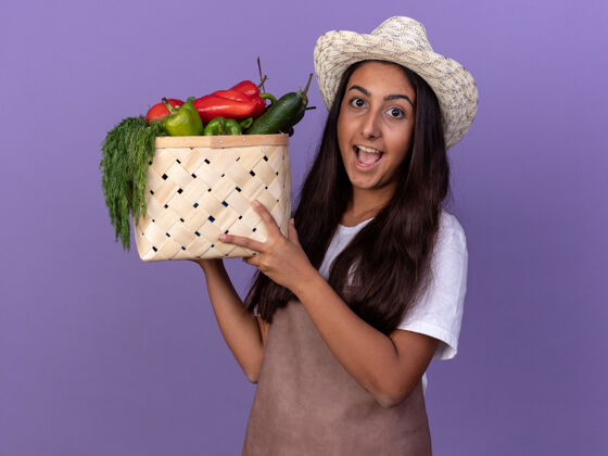 蔬菜穿着围裙 戴着夏帽的年轻园丁女孩站在紫色的墙上 手里拿着装满蔬菜的箱子 脸上洋溢着幸福的表情围裙幼嫩的帽子