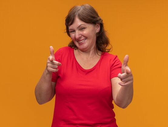 手指身穿红色t恤的中年妇女用食指指着橙色的墙上 快乐而愉快地站着目录中间站