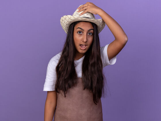 帽子穿着围裙 戴着夏令帽的年轻园丁女孩惊讶地碰了碰站在紫色墙上的帽子花园站立惊喜