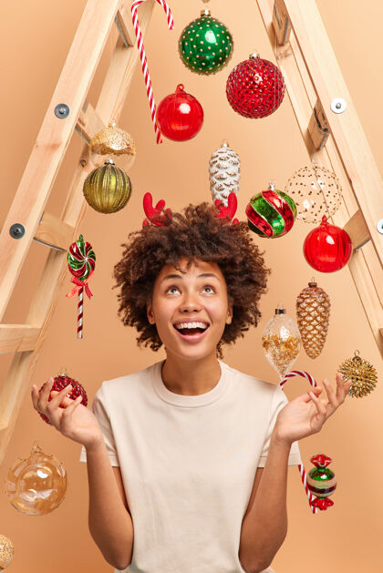 心情室内拍摄的快乐的黑皮肤女人高兴地看着上面摊开手掌和笑容广泛穿着红驯鹿角用梯子来装饰圣诞玩具周围的房子新年年玩具手掌